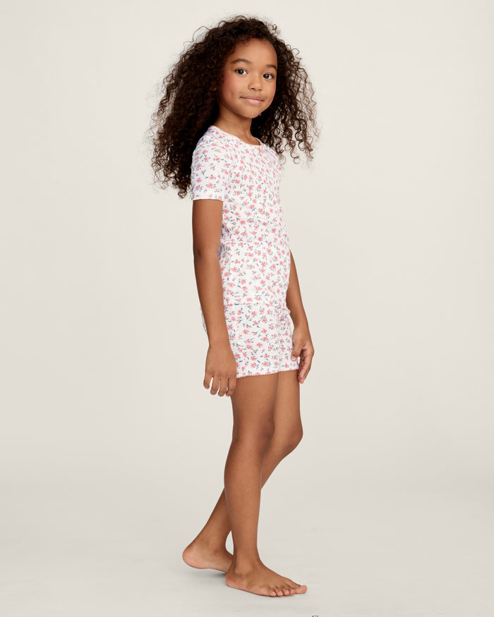 A young girl with curly hair stands against a plain background, wearing the Kid's Pointelle Snug Fit Pajama Short Set in Pink Floral, featuring a matching short-sleeve top and shorts decorated with a pink and red floral pattern.