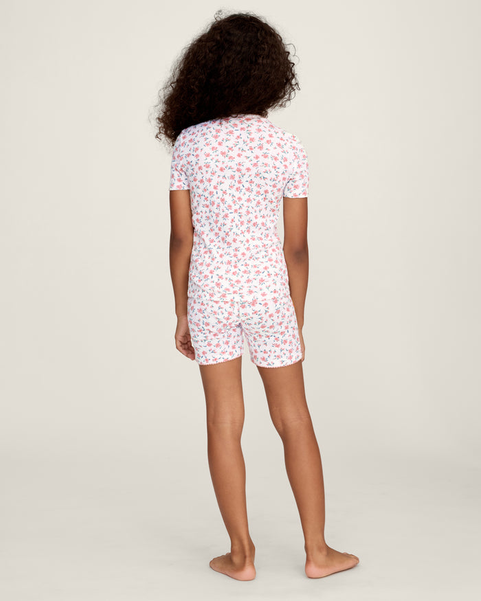 A young girl with curly hair stands against a plain background, wearing the Kid's Pointelle Snug Fit Pajama Short Set in Pink Floral, featuring a matching short-sleeve top and shorts decorated with a pink and red floral pattern.