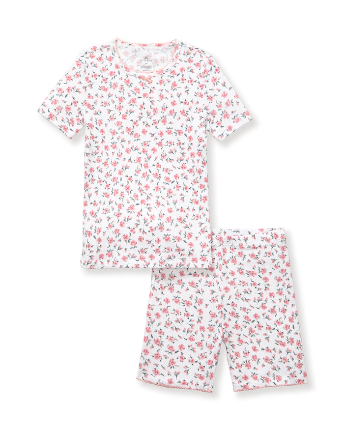 A young girl with curly hair stands against a plain background, wearing the Kid's Pointelle Snug Fit Pajama Short Set in Pink Floral, featuring a matching short-sleeve top and shorts decorated with a pink and red floral pattern.