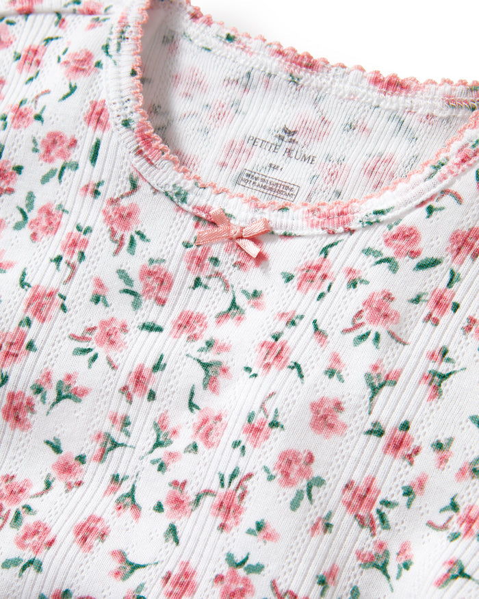 A young girl with curly hair stands against a plain background, wearing the Kid's Pointelle Snug Fit Pajama Short Set in Pink Floral, featuring a matching short-sleeve top and shorts decorated with a pink and red floral pattern.