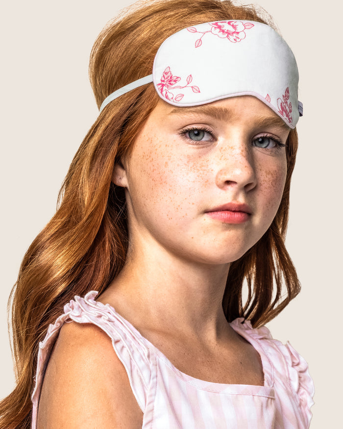 A young girl with long red hair gazes forward, wearing a Kids Sleep Mask in English Rose Floral by Petite Plume, with a pink sleeveless top against a neutral background. Perhaps this is the start of a memorable moment, just before gifting her matching eye masks to friends.