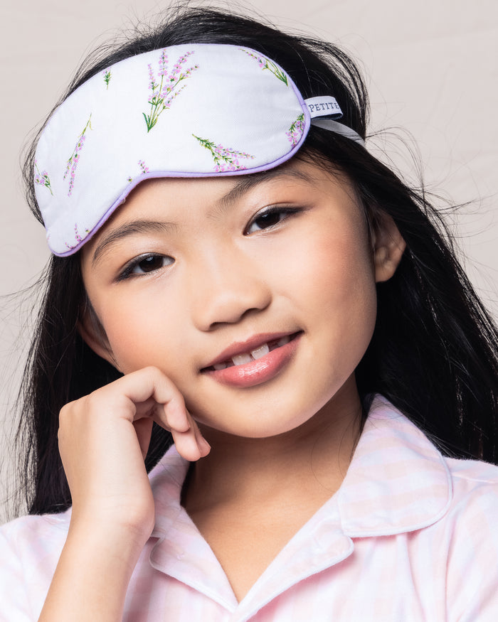 A young girl with long black hair smiles softly, wearing the Kids Sleep Mask in Fields of Provence by Petite Plume. This luxury piece complements her light pink plaid pajama top as her hand gently touches her cheek.