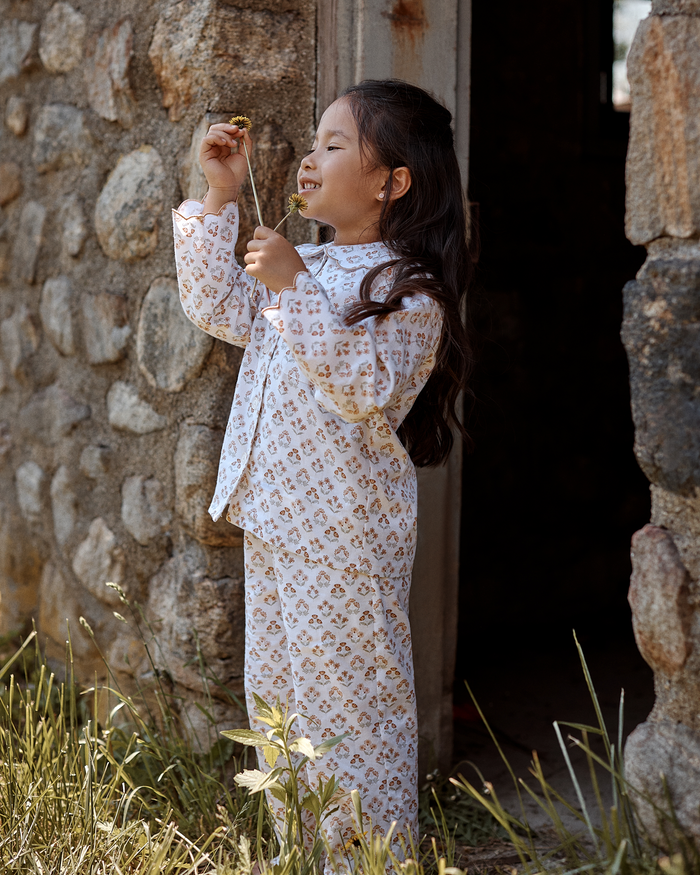 Wearing the Kid’s Twill Colette Pajama Set in Estate Garden, a young girl smiles as she stands by a stone wall, holding a yellow flower to her nose. She enjoys the sunshine outdoors amidst tall grass.