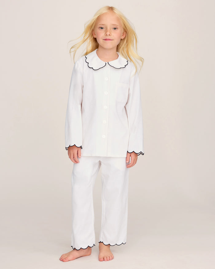 A young child with long blonde hair wears the Kid’s Twill Colette Pajama Set in White with Navy Piping, standing barefoot on a light background and looking forward with a gentle expression.