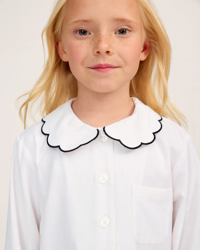 A young child with long blonde hair is wearing the Kid’s Twill Colette Pajama Set in White with Navy Piping, featuring a white buttoned shirt with a scalloped collar trimmed in navy. The child is facing forward and smiling slightly.