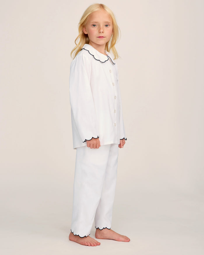 A young child with long blonde hair stands barefoot, wearing the Kid’s Twill Colette Pajama Set in White with Navy Piping. The pajamas feature navy piping along the collar, cuffs, and pant hems. The background is plain and light-colored.