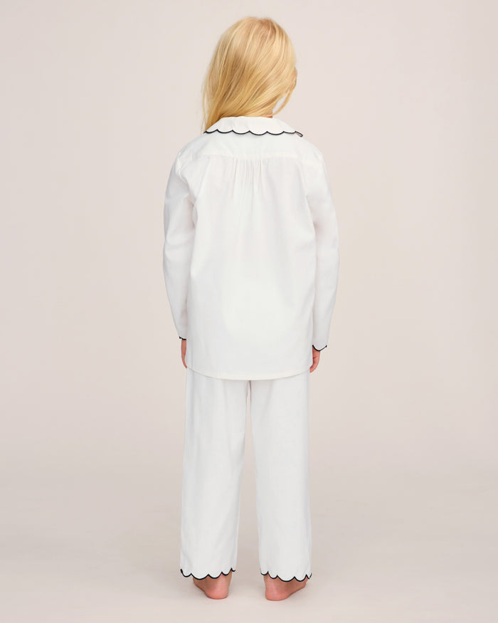 A young child with long blonde hair stands barefoot, facing away, wearing the Kid’s Twill Colette Pajama Set in White with Navy Piping against a plain light background.