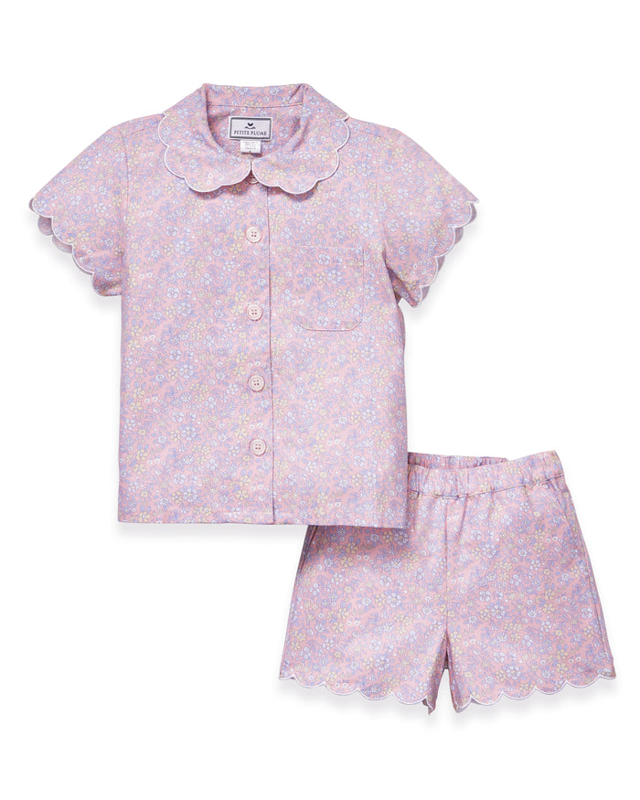 A young girl with long blonde hair stands facing forward, wearing the Kid's Twill Colette Short Set in Fleur Blush—an adorable pastel purple and pink pajama set with scalloped edges—against a plain light background.