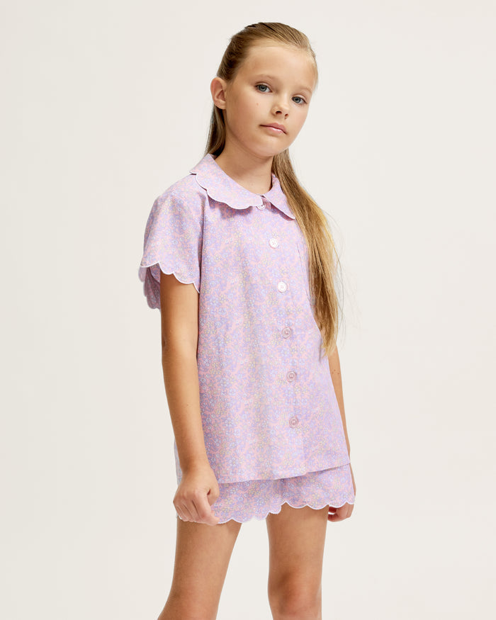 A young girl with long blonde hair stands facing forward, wearing the Kid's Twill Colette Short Set in Fleur Blush—an adorable pastel purple and pink pajama set with scalloped edges—against a plain light background.