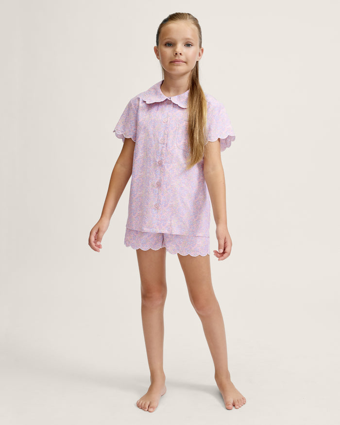 A young girl with long blonde hair stands facing forward, wearing the Kid's Twill Colette Short Set in Fleur Blush—an adorable pastel purple and pink pajama set with scalloped edges—against a plain light background.