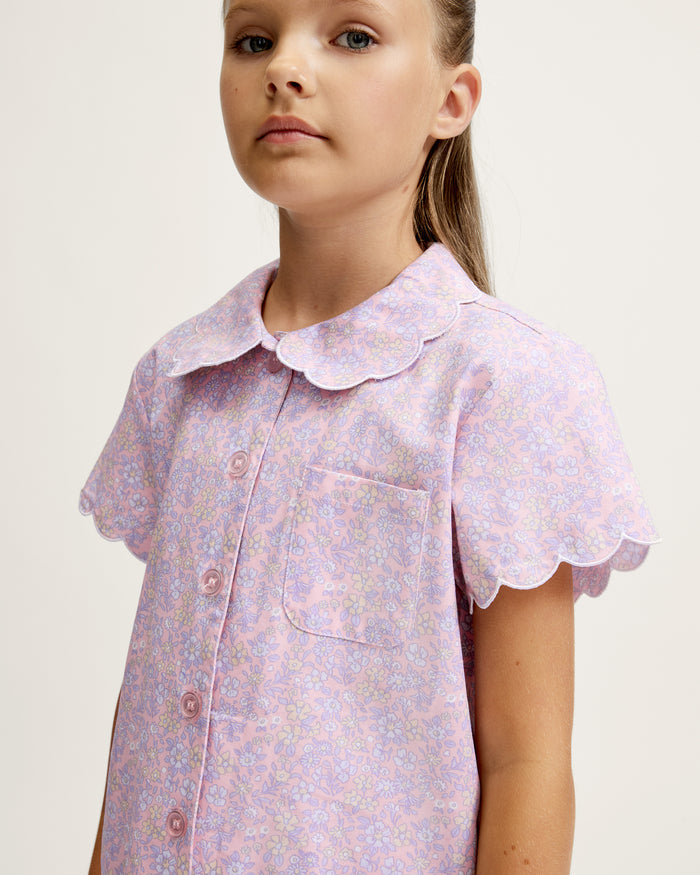 A young girl with long blonde hair stands facing forward, wearing the Kid's Twill Colette Short Set in Fleur Blush—an adorable pastel purple and pink pajama set with scalloped edges—against a plain light background.