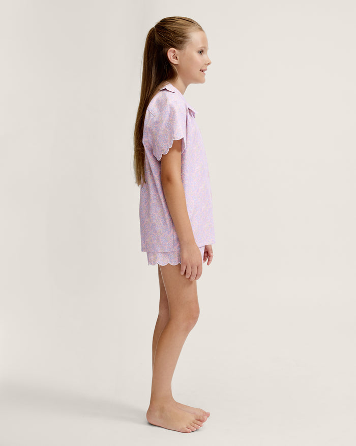 A young girl with long blonde hair stands facing forward, wearing the Kid's Twill Colette Short Set in Fleur Blush—an adorable pastel purple and pink pajama set with scalloped edges—against a plain light background.