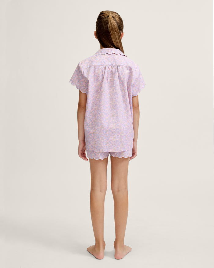 A young girl with long blonde hair stands facing forward, wearing the Kid's Twill Colette Short Set in Fleur Blush—an adorable pastel purple and pink pajama set with scalloped edges—against a plain light background.
