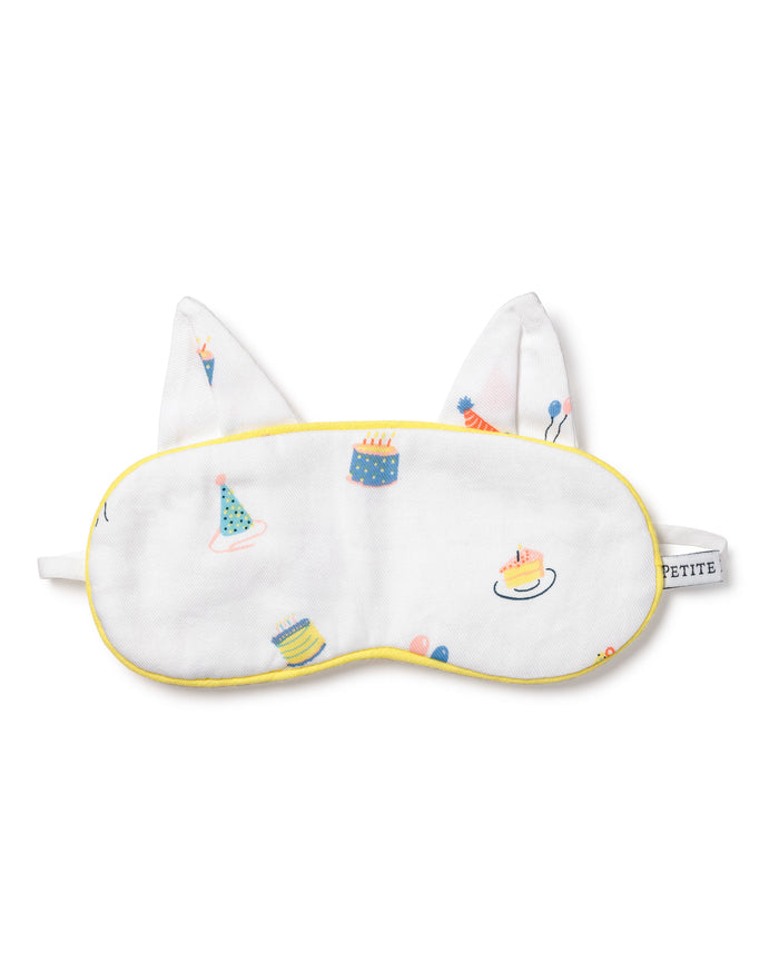 The Kids Twill Kitty Sleep Mask in Birthday Wishes by Petite Plume is a luxurious white eye mask with yellow trim, featuring colorful cake, party hat, and balloon illustrations. It includes two fabric ears, an elastic strap, and can be monogrammed for personalization.