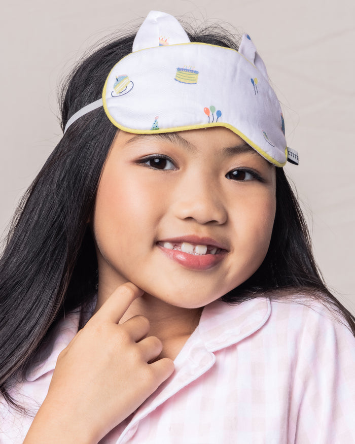 A young girl with long dark hair smiles wearing the Kids Twill Kitty Sleep Mask in Birthday Wishes by Petite Plume. She is dressed in a pink checkered pajama top and has her hand gently resting under her chin.