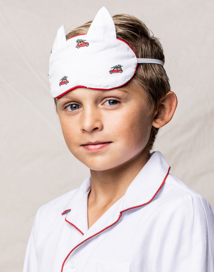 A young boy, in luxurious sleepwear from Petite Plume, wears the Kids Twill Kitty Sleep Mask in Holiday Journey with red car and tree designs. The mask perfectly matches his pajamas with red piping, adding charm against a soft light beige background.