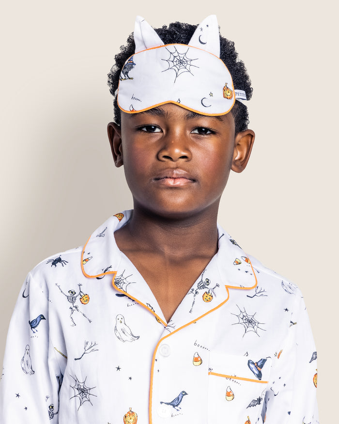 A child in luxurious Halloween-themed pajamas features Trick or Treat prints with pumpkins, ghosts, and birds, and wears a Petite Plume Kids Twill Kitty Sleep Mask with spiderweb designs and playful cat ears against a plain light gray background.