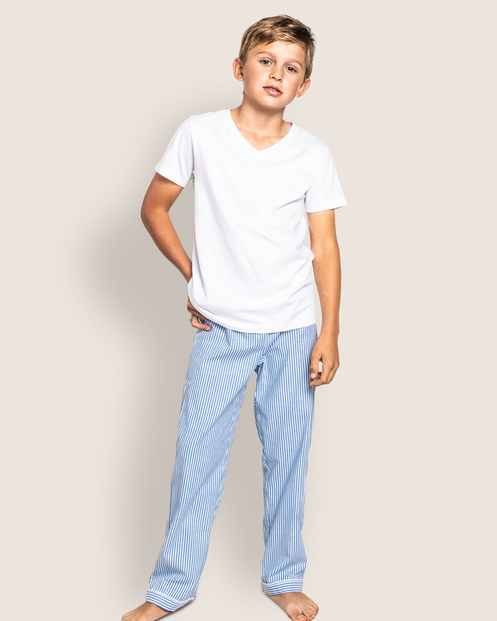 A young boy stands barefoot against a simple light background, dressed in luxurious sleepwear from Petite Plume. His white T-shirt pairs with Kids Twill Pajama Pants in French Blue Seersucker, crafted from fine yarn-dyed cotton. One hand rests in a pocket, enhancing the serene scene.