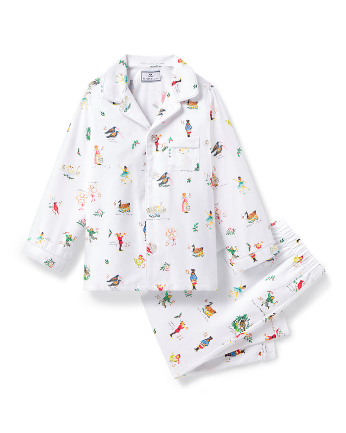 The Kid's Twill Pajama Set in 12 Days of Christmas features a white, long-sleeve button-up top with collar and front pocket, plus matching pants, all adorned with playful, colorful holiday illustrations of people and animals.