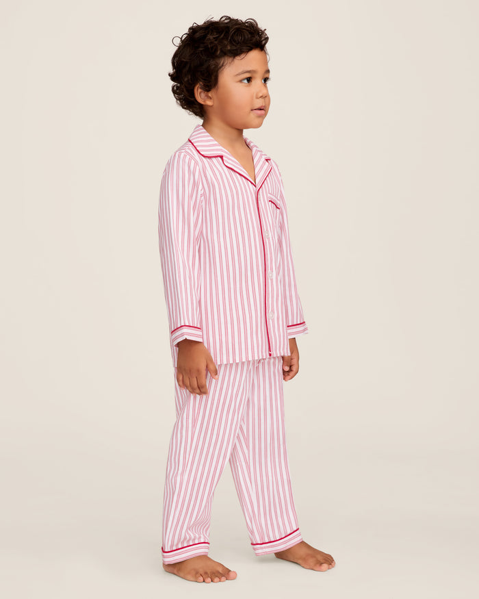 A man and a child sit on a wooden window seat, wearing matching yarn-dyed cotton pajamas. The man wears a pink striped set, while the child dons Petite Plumes Kids Twill Pajama Set in Antique Red Ticking, paired with a red robe. They are reading together.