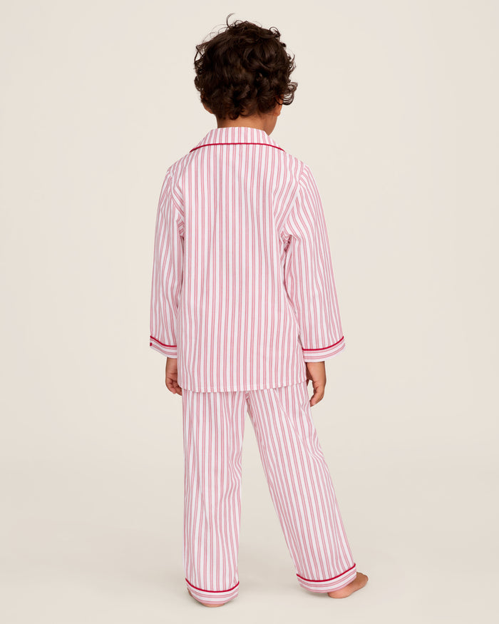 A man and a child sit on a wooden window seat, wearing matching yarn-dyed cotton pajamas. The man wears a pink striped set, while the child dons Petite Plumes Kids Twill Pajama Set in Antique Red Ticking, paired with a red robe. They are reading together.