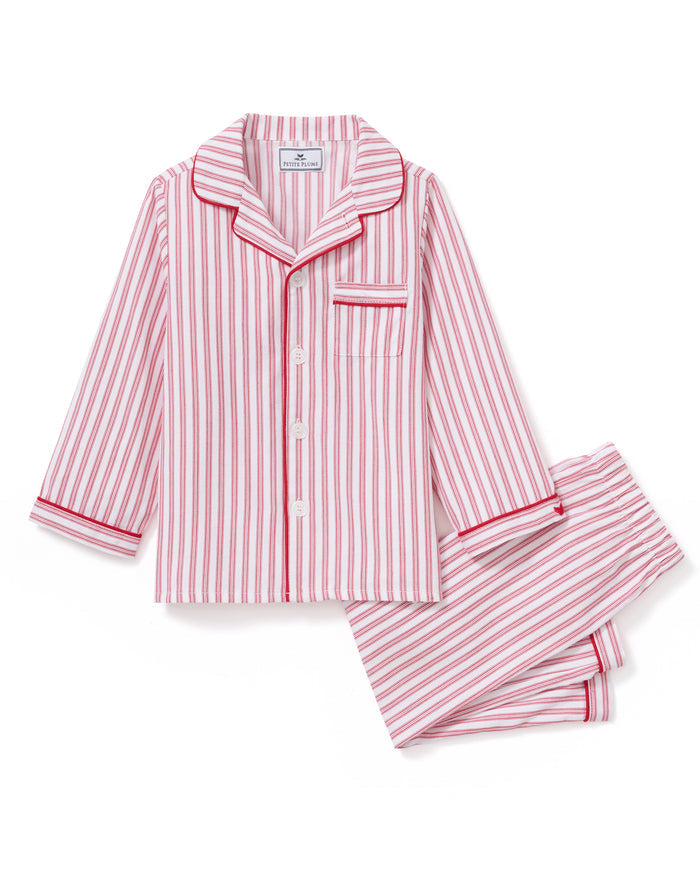 A man and a child sit on a wooden window seat, wearing matching yarn-dyed cotton pajamas. The man wears a pink striped set, while the child dons Petite Plumes Kids Twill Pajama Set in Antique Red Ticking, paired with a red robe. They are reading together.