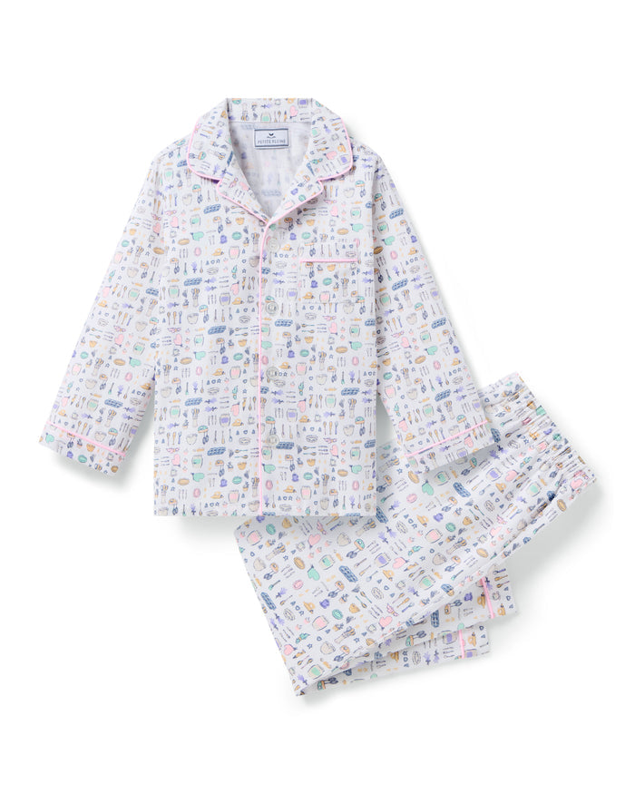 The Kid's Twill Pajama Set in Baking Spirits Bright features white long-sleeve button-up top and long pants with a colorful print of various small household and travel items.