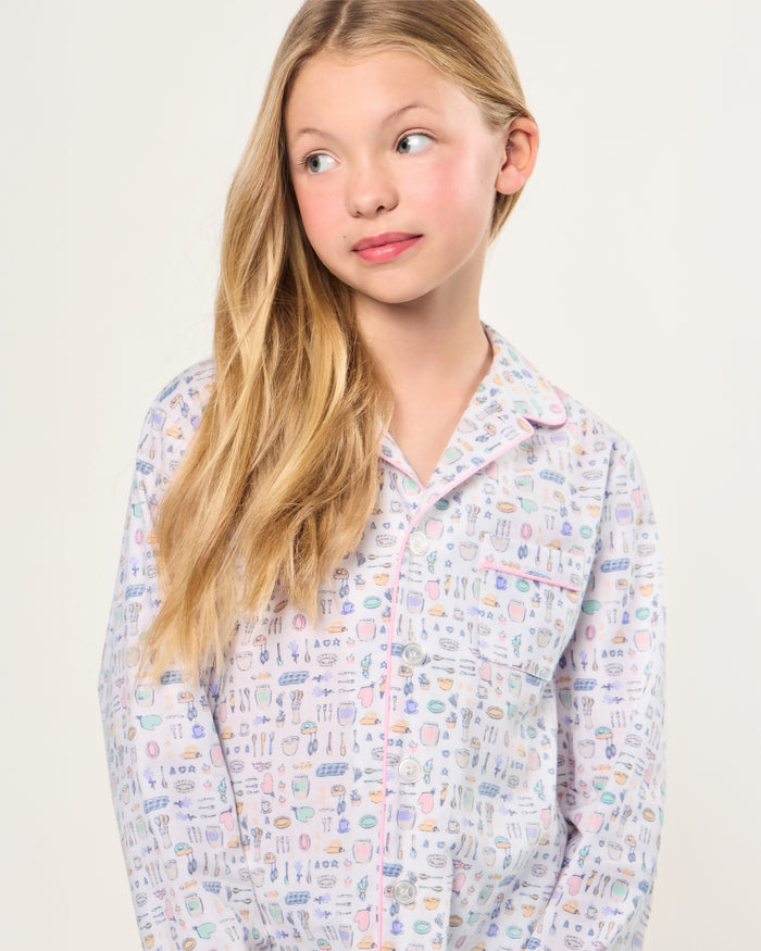 The Kid's Twill Pajama Set in Baking Spirits Bright features white long-sleeve button-up top and long pants with a colorful print of various small household and travel items.