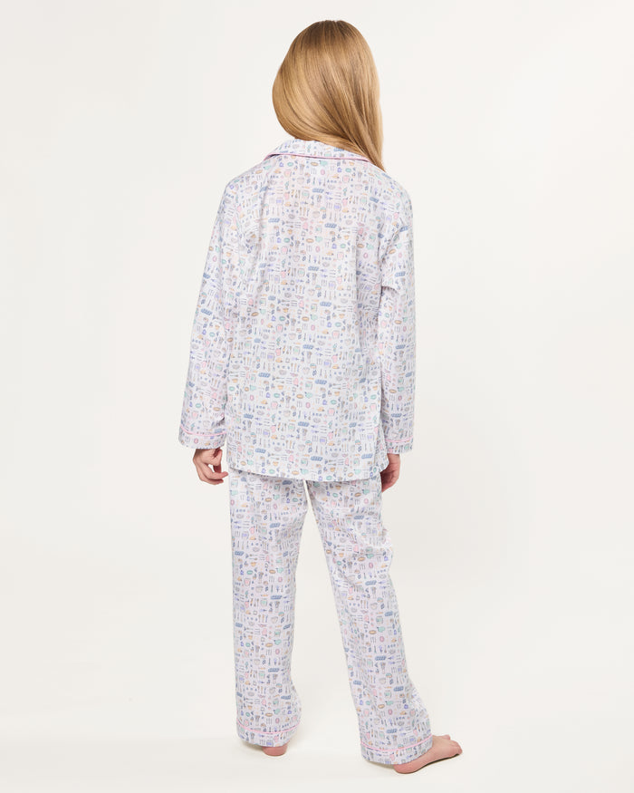 The Kid's Twill Pajama Set in Baking Spirits Bright features white long-sleeve button-up top and long pants with a colorful print of various small household and travel items.