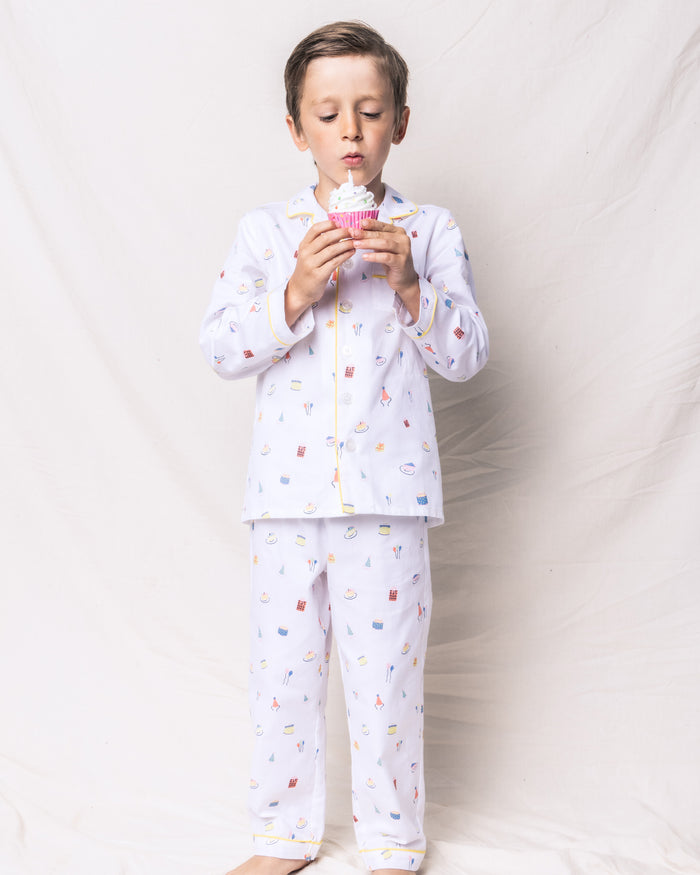A child in Petite Plumes Kids Twill Pajama Set in Birthday Wishes stands against a light background, holding a white-frosted cupcake. The luxurious sleepwear features small images with yellow piping, creating a charming birthday fantasy print.