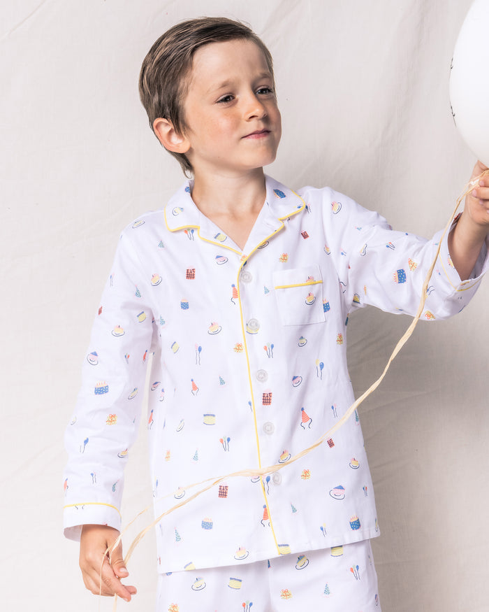 A young child with short hair wears Petite Plumes Kids Twill Pajama Set in Birthday Wishes, showcasing dessert and utensil designs. Holding a balloon string, the child stands against a plain white background.