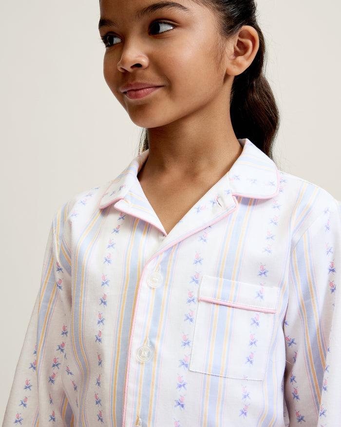 A young girl with dark hair stands barefoot on a plain background, wearing the Kid's Twill Pajama Set in Cottage Stripe—matching white pajamas with pastel stripes and a chest pocket—facing forward and smiling slightly.
