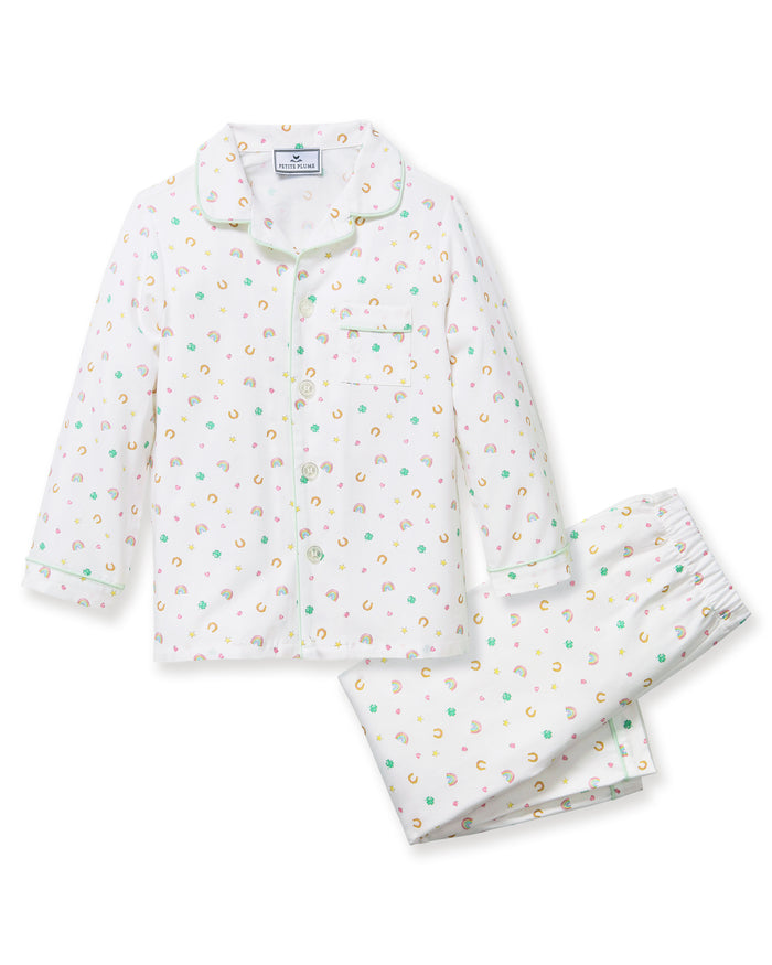 A child with short curly hair stands facing forward, wearing the Kid's Twill Pajama Set in Good Luck Charms—a white button-up pajama with a colorful pattern and matching pants—against a plain light background.