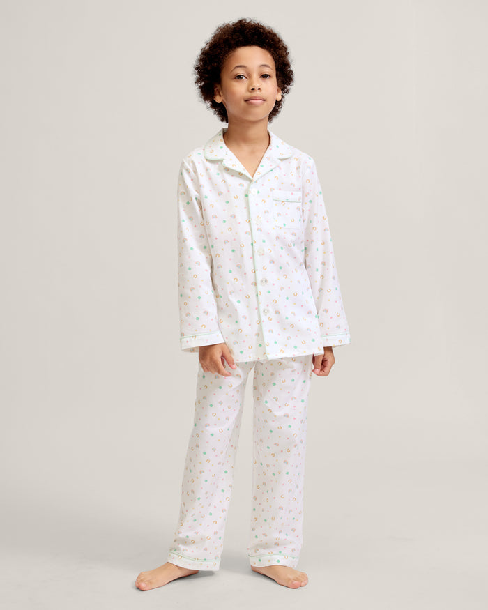 A child with short curly hair stands facing forward, wearing the Kid's Twill Pajama Set in Good Luck Charms—a white button-up pajama with a colorful pattern and matching pants—against a plain light background.