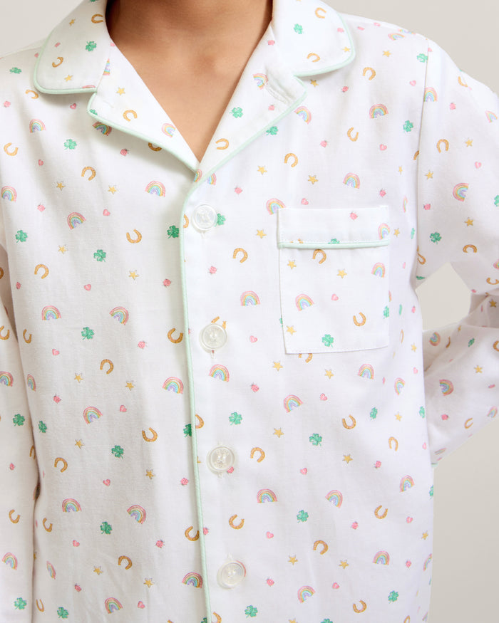 A child with short curly hair stands facing forward, wearing the Kid's Twill Pajama Set in Good Luck Charms—a white button-up pajama with a colorful pattern and matching pants—against a plain light background.