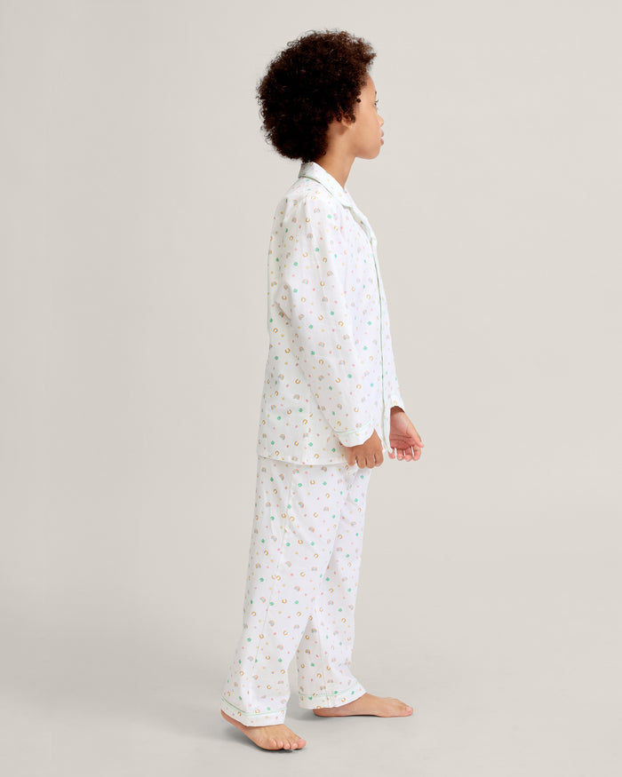 A child with short curly hair stands facing forward, wearing the Kid's Twill Pajama Set in Good Luck Charms—a white button-up pajama with a colorful pattern and matching pants—against a plain light background.