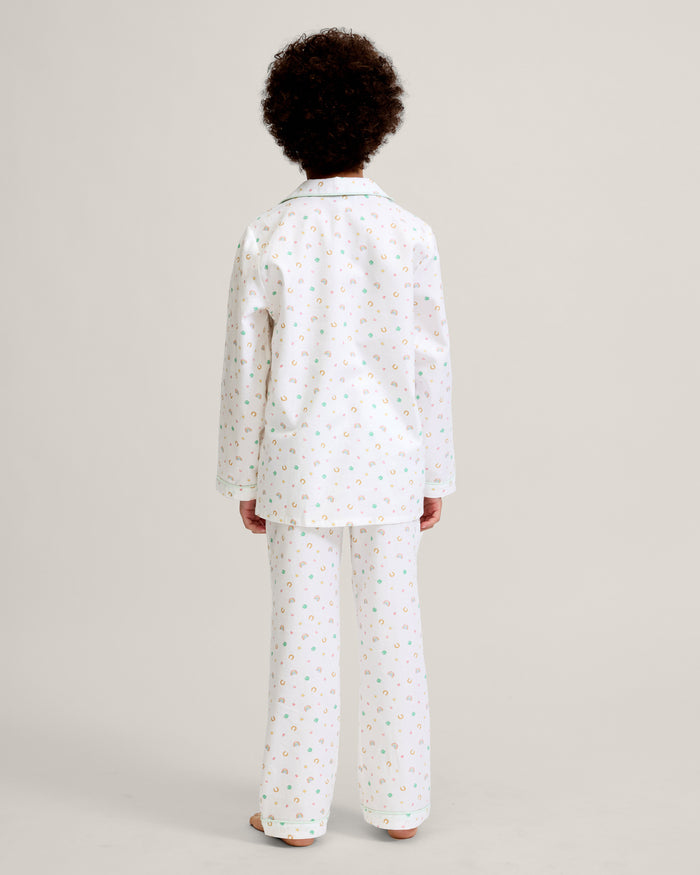 A child with short curly hair stands facing forward, wearing the Kid's Twill Pajama Set in Good Luck Charms—a white button-up pajama with a colorful pattern and matching pants—against a plain light background.