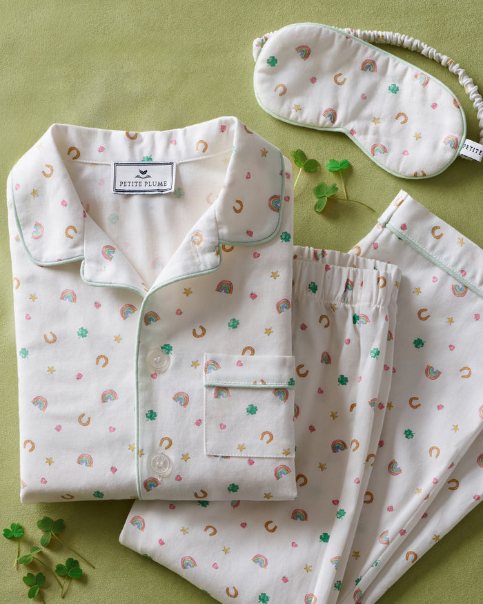 A child with short curly hair stands facing forward, wearing the Kid's Twill Pajama Set in Good Luck Charms—a white button-up pajama with a colorful pattern and matching pants—against a plain light background.