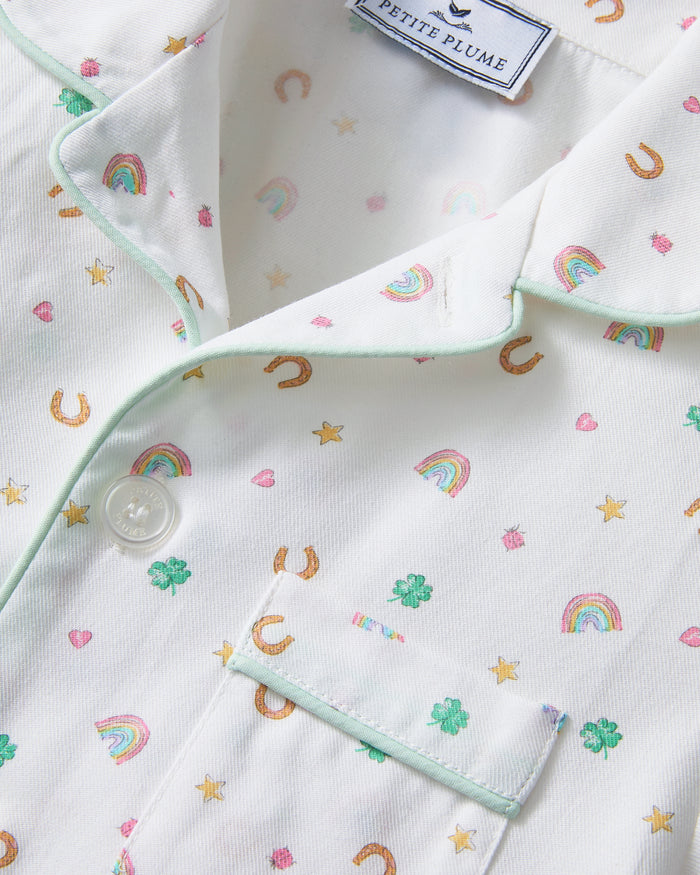 A child with short curly hair stands facing forward, wearing the Kid's Twill Pajama Set in Good Luck Charms—a white button-up pajama with a colorful pattern and matching pants—against a plain light background.