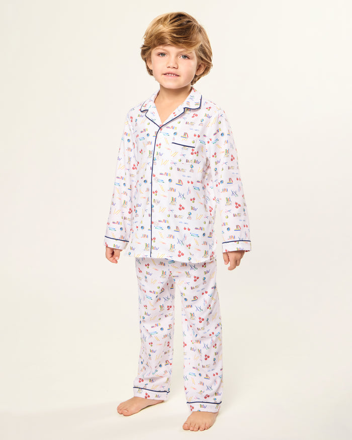 The Kid's Twill Pajama Set in Literary Lessons features a white collared, long-sleeve button-up top and matching pants with a colorful school-themed pattern and navy blue piping accents.