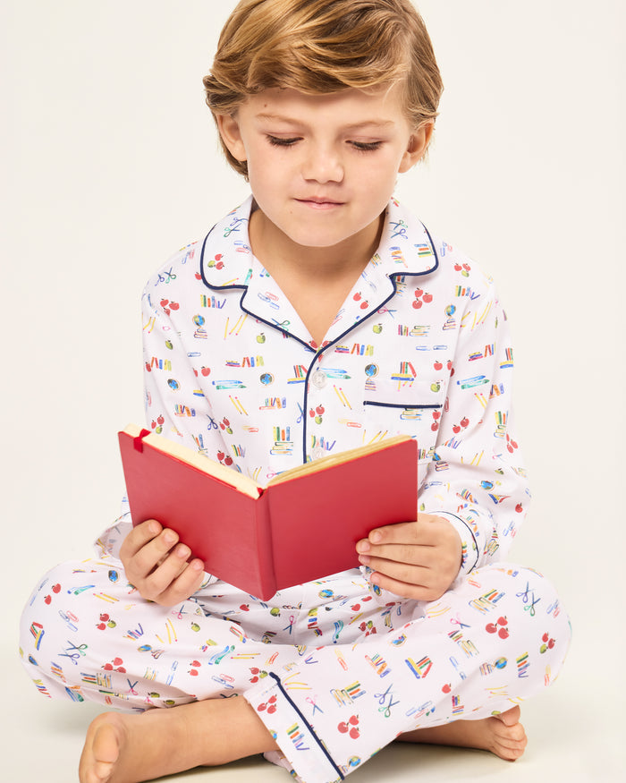 The Kid's Twill Pajama Set in Literary Lessons features a white collared, long-sleeve button-up top and matching pants with a colorful school-themed pattern and navy blue piping accents.