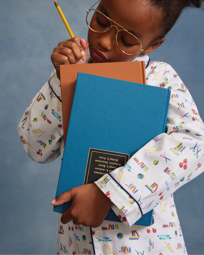 The Kid's Twill Pajama Set in Literary Lessons features a white collared, long-sleeve button-up top and matching pants with a colorful school-themed pattern and navy blue piping accents.