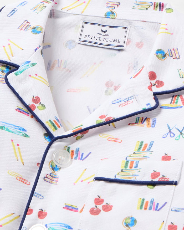 The Kid's Twill Pajama Set in Literary Lessons features a white collared, long-sleeve button-up top and matching pants with a colorful school-themed pattern and navy blue piping accents.