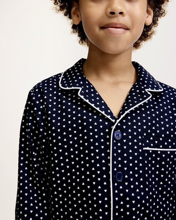 Kid's Twill Pajama Set in Midnight Hearts features navy blue pajamas with white heart prints, white piping, a long-sleeve button-up top with chest pocket, and matching pants.