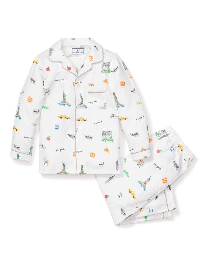 The Kids Twill Pajama Set in New York! New York! by Petite Plume features white pajamas with colorful prints of the Statue of Liberty, Empire State Building, taxis, and Brooklyn Bridge. The set includes a button-up shirt and pants adorned with pizza slices, pretzels, and New York script.