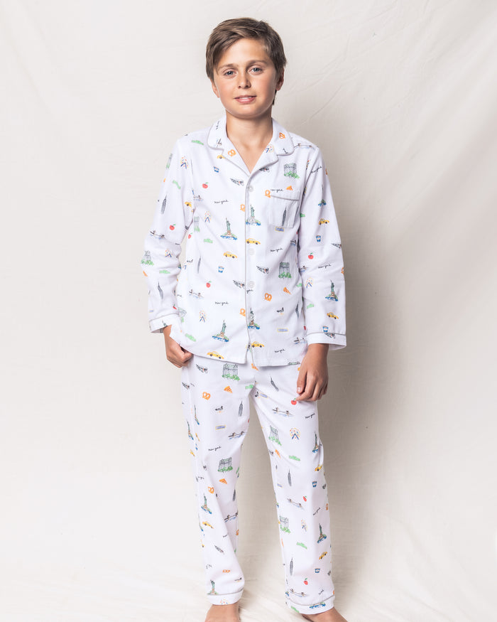 A young boy with short brown hair stands in front of a light background, wearing the Petite Plume Kids Twill Pajama Set in New York! New York! featuring vibrant cartoon patterns, including animals and cars. He has a relaxed expression and one hand in his pocket.
