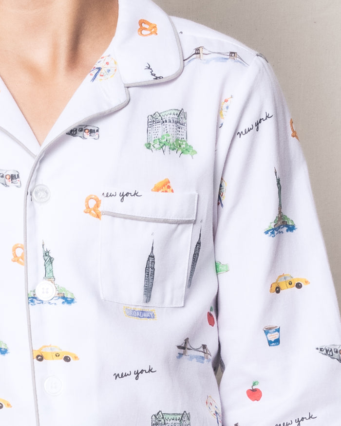 A kid is wearing Petite Plumes luxurious Twill Pajama Set in New York! New York!, featuring iconic designs like the Statue of Liberty, Brooklyn Bridge, pretzels, and yellow taxis. This classic set includes a pocket and front buttons.