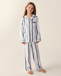 A young girl with long hair stands barefoot on a neutral background, slightly smiling with her head tilted, wearing the Kid’s Twill Pajama Set in Parlor Stripe—white pajamas featuring blue vertical stripes.