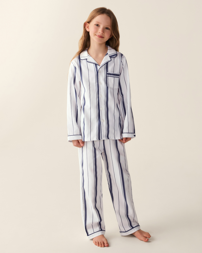 A young girl with long hair stands barefoot on a neutral background, slightly smiling with her head tilted, wearing the Kid’s Twill Pajama Set in Parlor Stripe—white pajamas featuring blue vertical stripes.