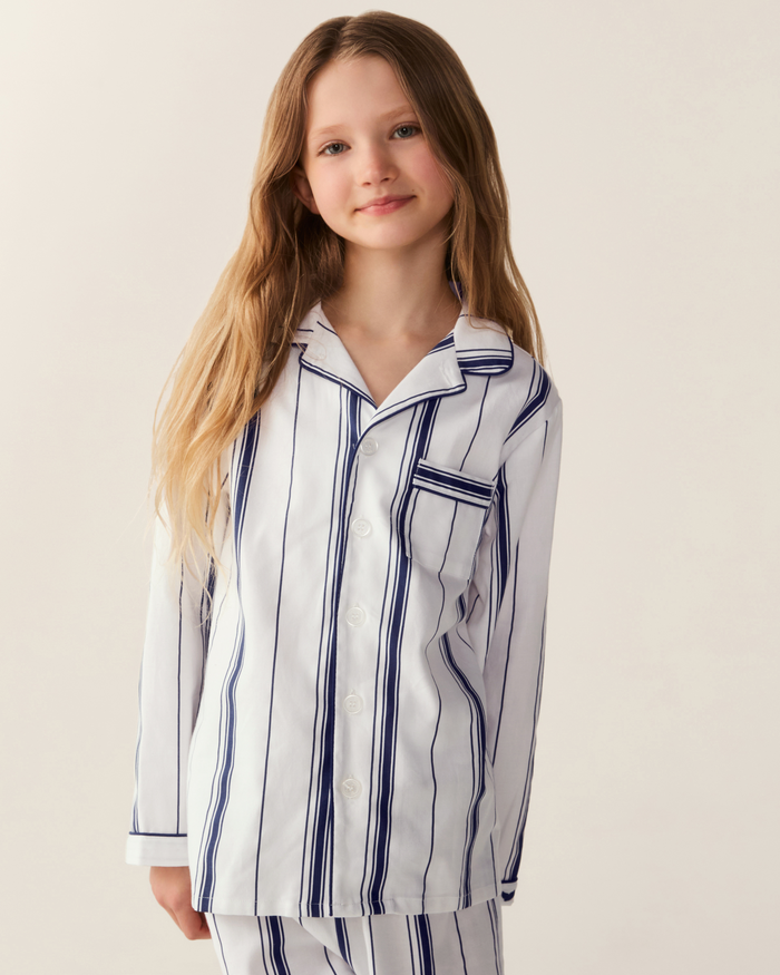 A young girl with long, light brown hair wears the Kid’s Twill Pajama Set in Parlor Stripe—featuring white fabric with navy blue vertical stripes—standing against a plain, light background and smiling gently at the camera.